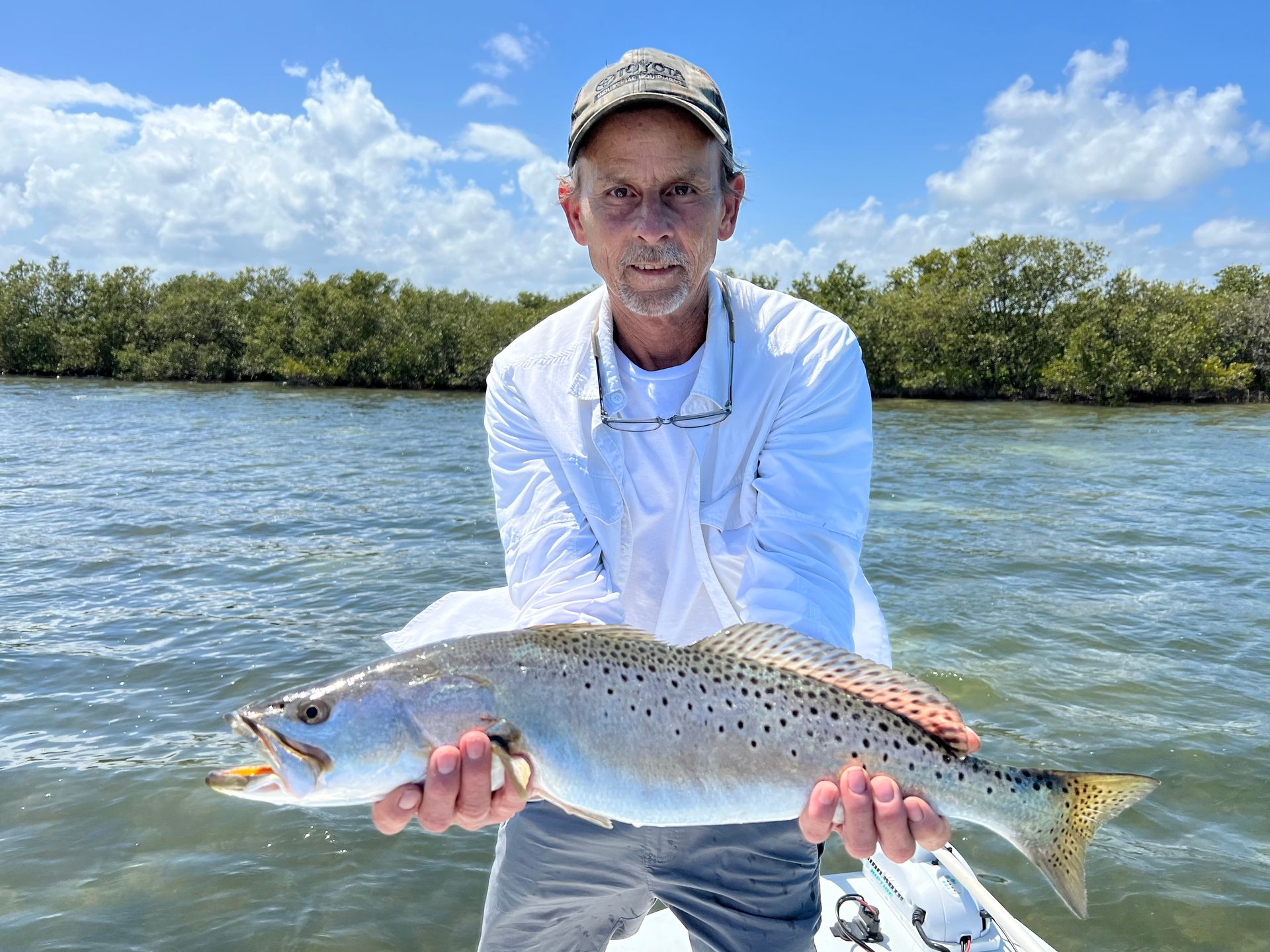 Man on a boat, smiling, holding a blue and green fish he caught. Ocean background, sunny day.