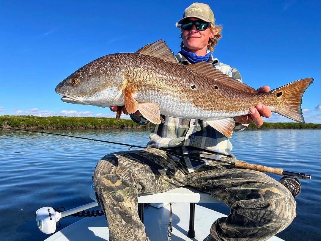 Man in camouflage holds a large redfish on a boat in a marsh, sunny day.
