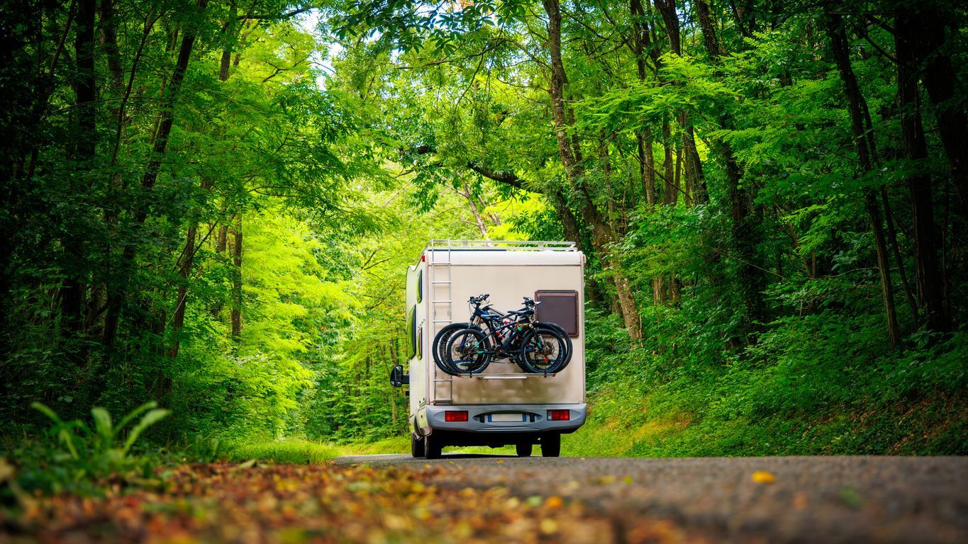 A rv with two bikes on the back is driving down a road in the woods.
