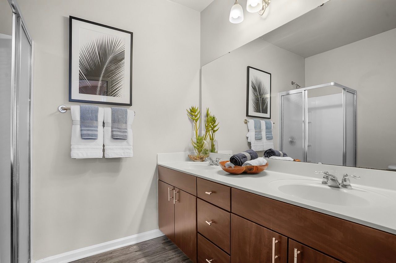 Bathroom with double vanity, wood cabinets, large mirror, framed art, towels, and glass shower.