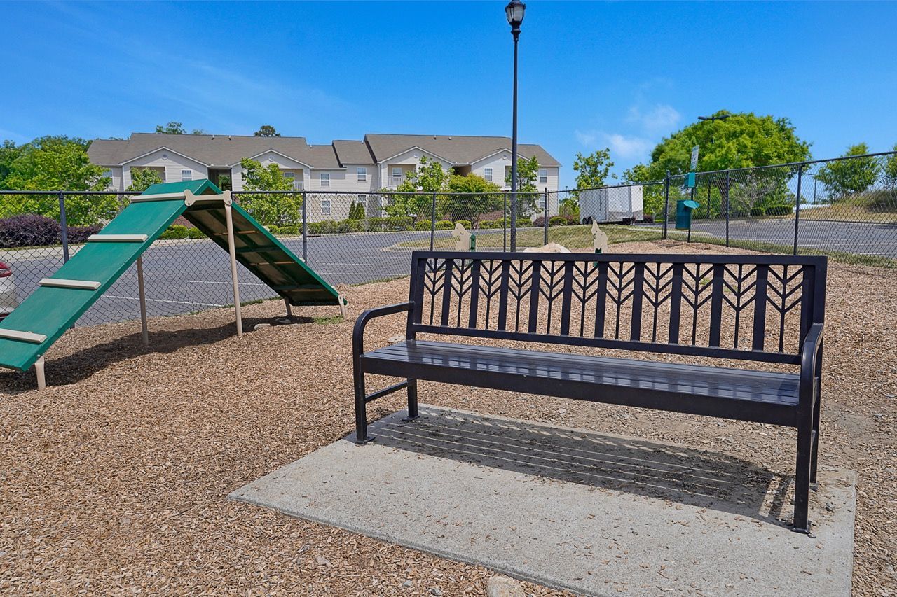Metal bench on a concrete pad beside a green playground, with apartments in the background.