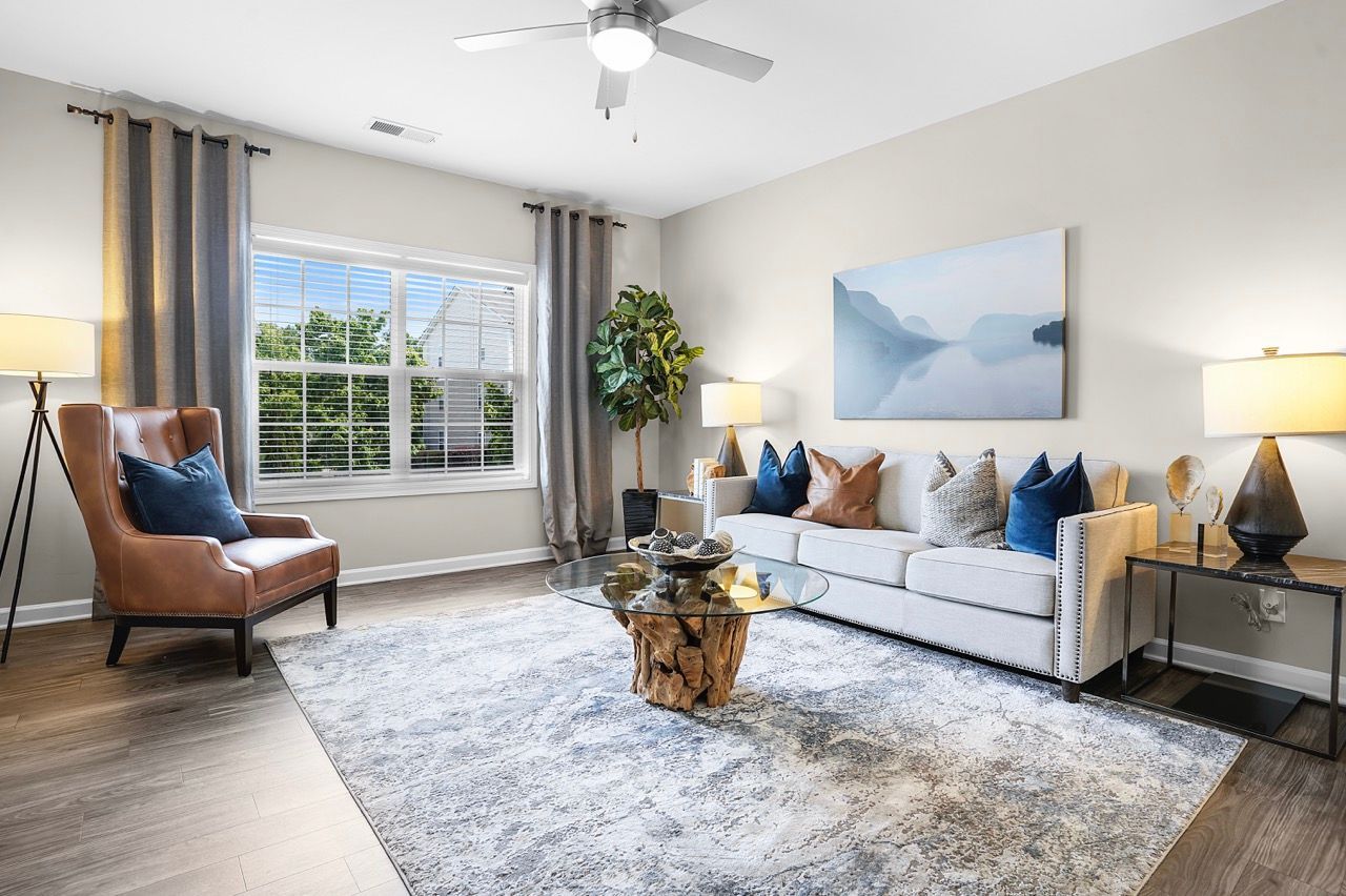 Bright living room with gray sofa, brown armchair, glass coffee table, and large window with curtains.