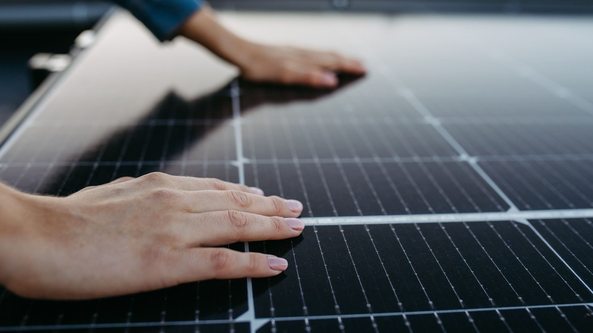 Hands of a person touching a solar panel, likely during installation. Close-up shows the dark, textured surface of the panel.