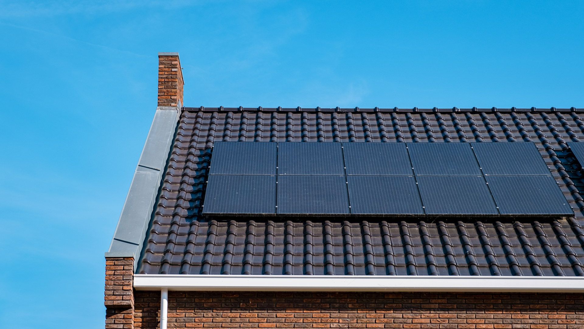 Solar panels on a brick house roof against a blue sky. The roof has dark tiles and a brick chimney.