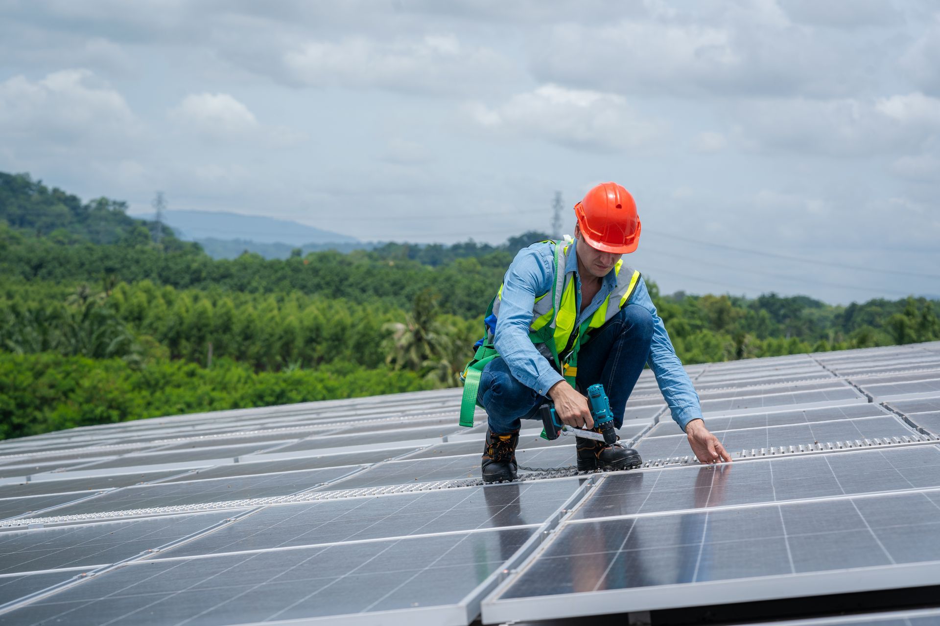 A worker in a hard hat and safety vest installs solar panels on a rooftop, surrounded by green trees under a cloudy sky.