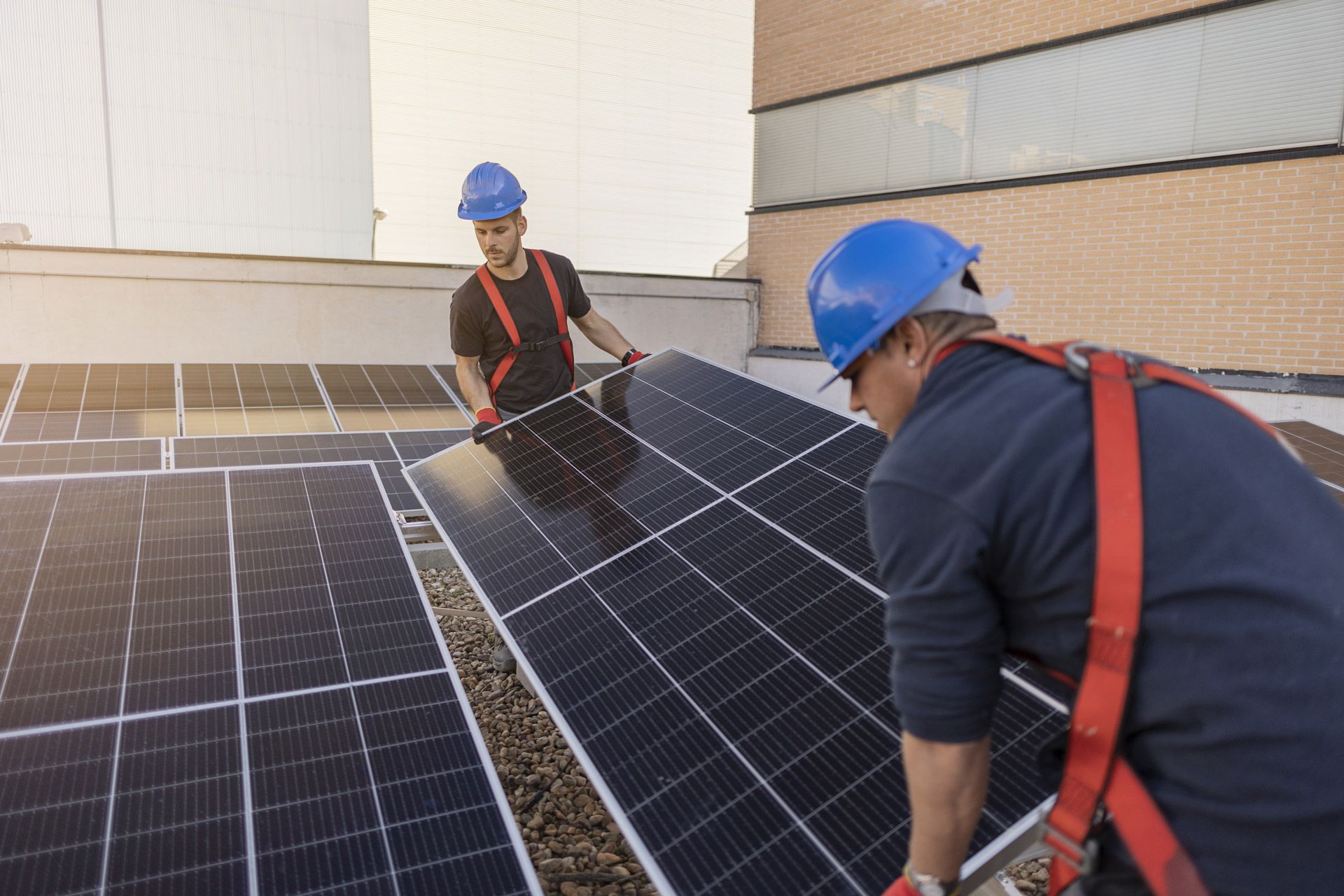 Two workers in hard hats install solar panels on a rooftop; they wear safety harnesses.