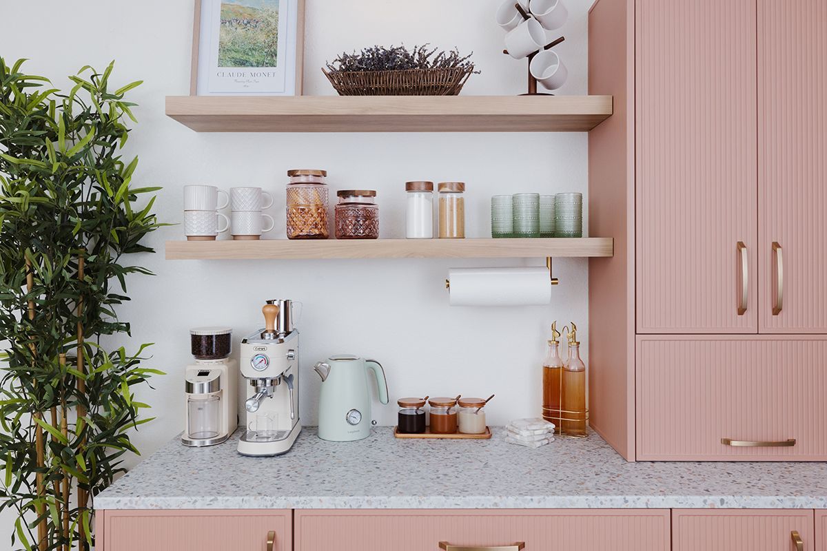 Pink kitchen with open shelves holding jars, glasses, and a paper towel roll. A green plant and a light blue kettle are present.