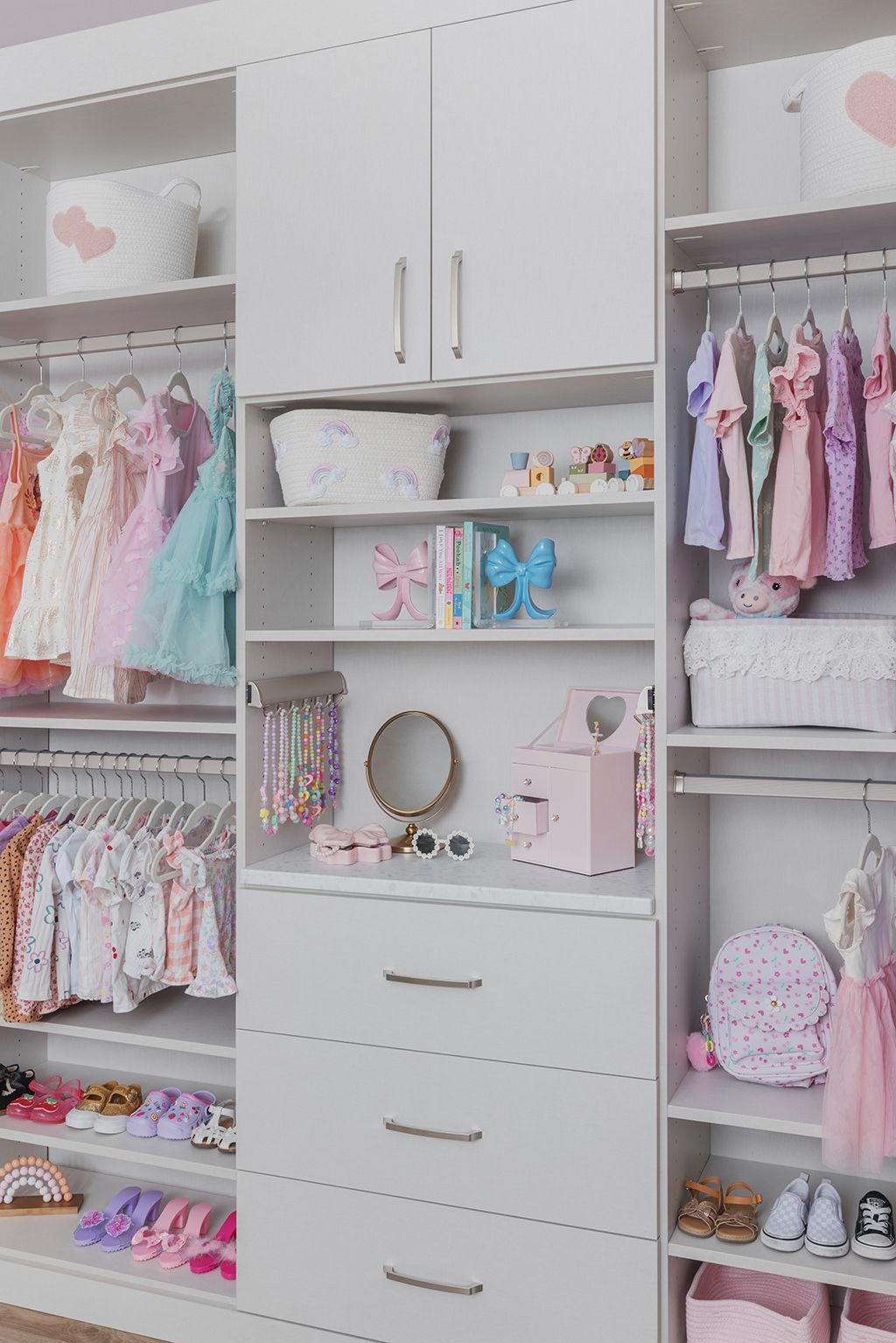 White closet with shelves, hanging pastel clothes, folded items, and drawers organized in a tidy children’s room.