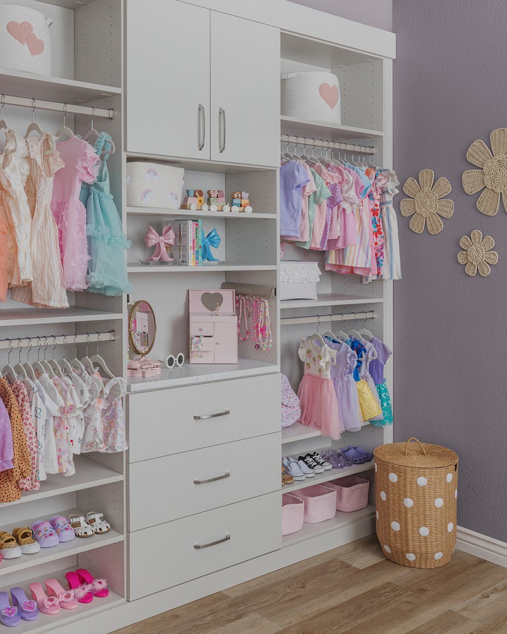 Children’s closet with pastel clothes, white shelving, drawers, and decorative wall flowers on a purple wall