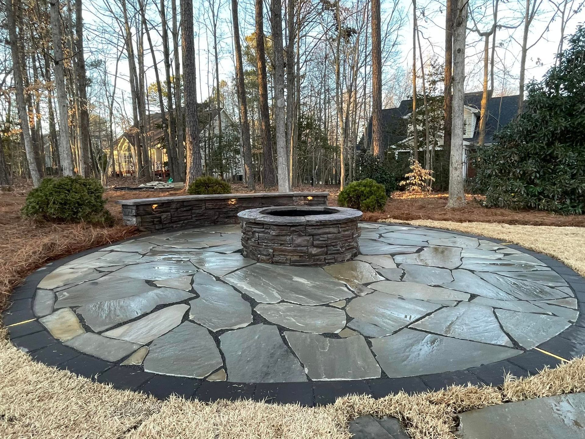 A stone patio with a fire pit in the middle of it surrounded by trees.
