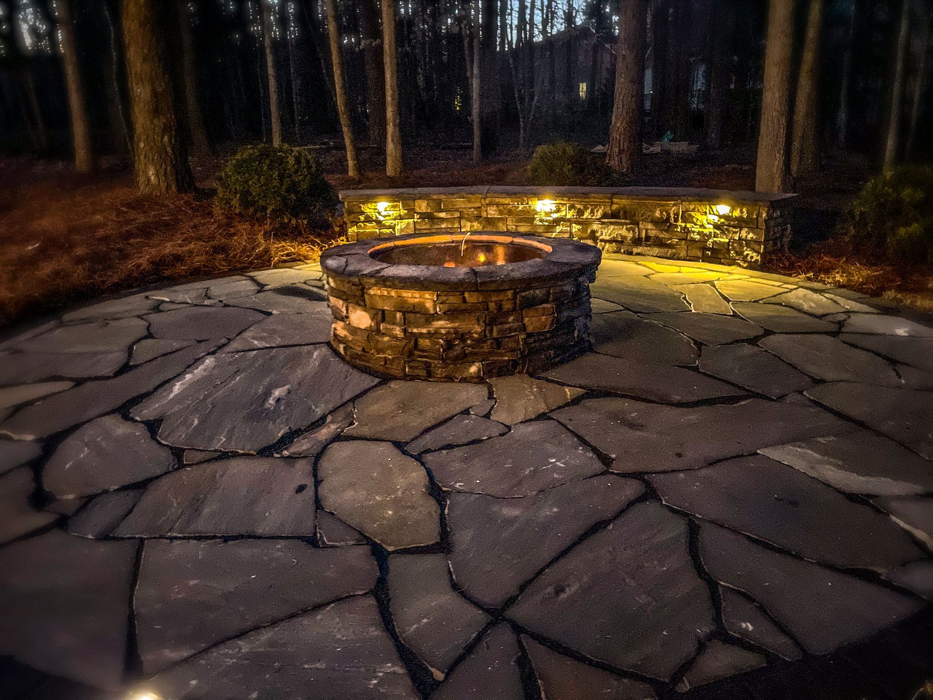 A fire pit in the middle of a stone patio in the woods.