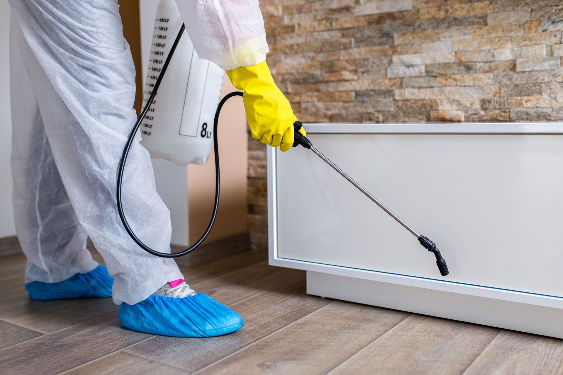 Person in protective gear spraying insecticide near a white cabinet and stone wall.