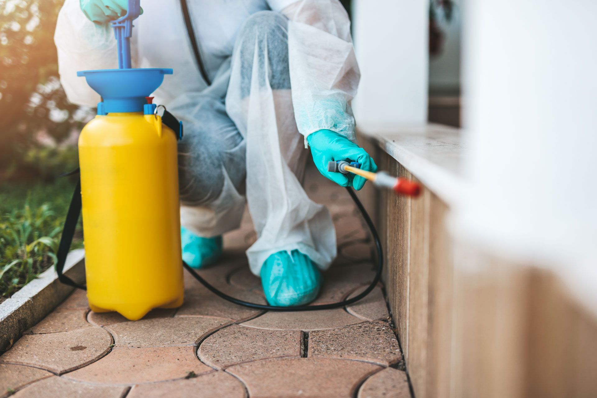 Person in protective suit spraying pesticide along a building foundation.
