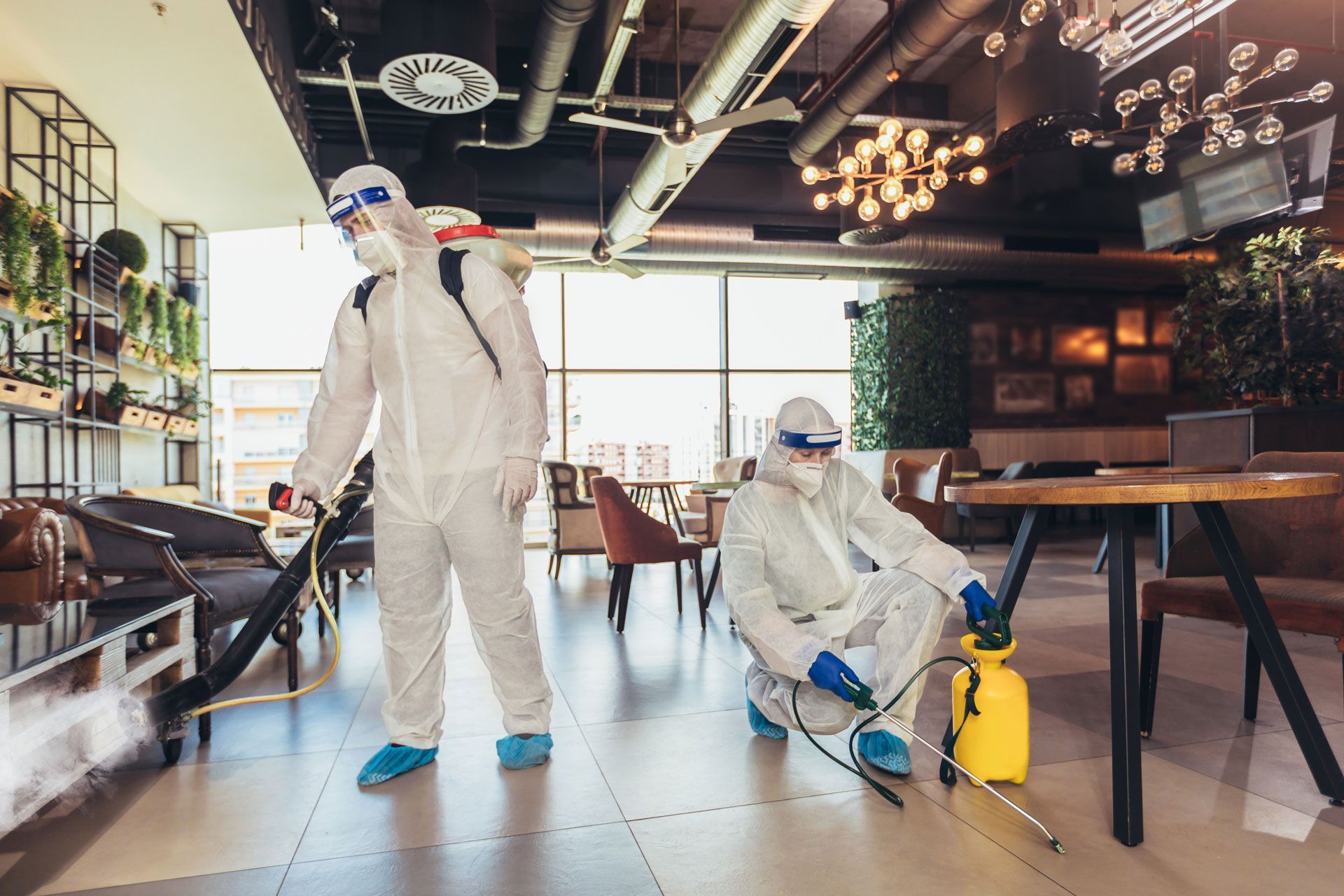 Two people in hazmat suits disinfecting a commercial interior with sprayers.
