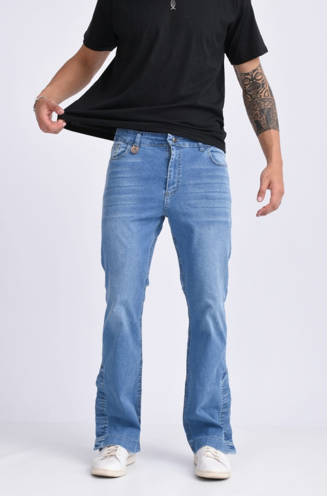 Persona con camiseta negra, vaqueros azul claro y zapatillas blancas de pie sobre un fondo liso.
