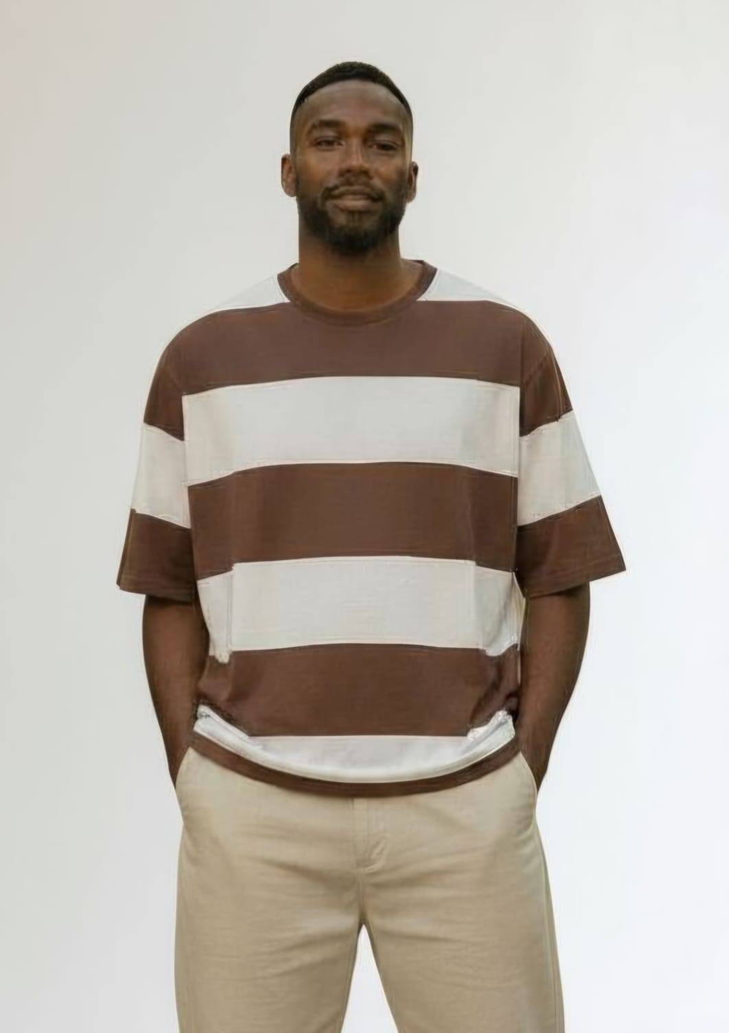 Persona de pie contra una pared blanca, vestida con una camisa de rayas marrones y blancas y pantalones beige.