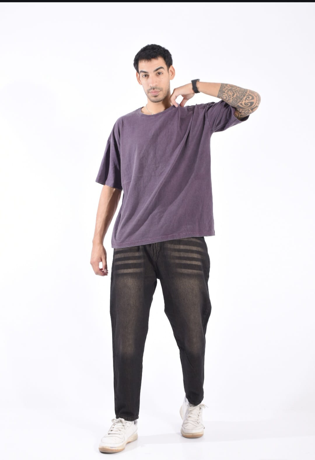Hombre con camiseta morada, pantalones negros y zapatillas blancas posando sobre un fondo blanco.
