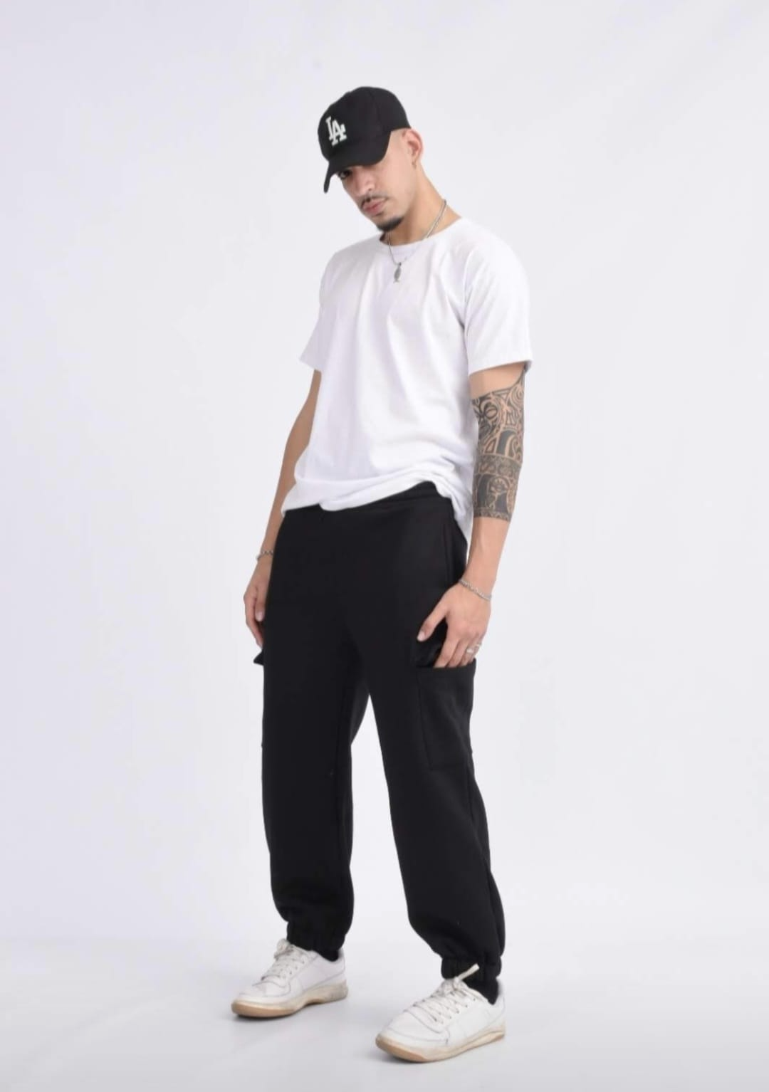 Hombre con camiseta blanca, pantalones negros, gorra y zapatillas blancas posando sobre un fondo gris claro.