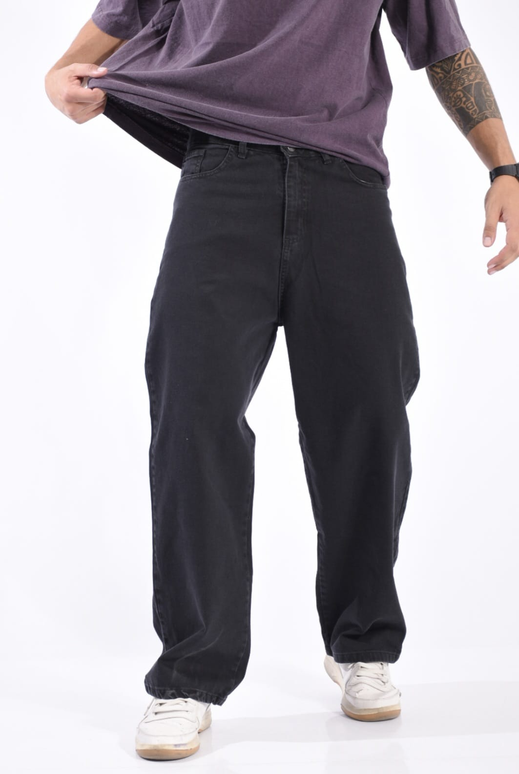 Persona con pantalones negros de pierna ancha, camisa morada y zapatillas blancas sobre un fondo blanco liso.