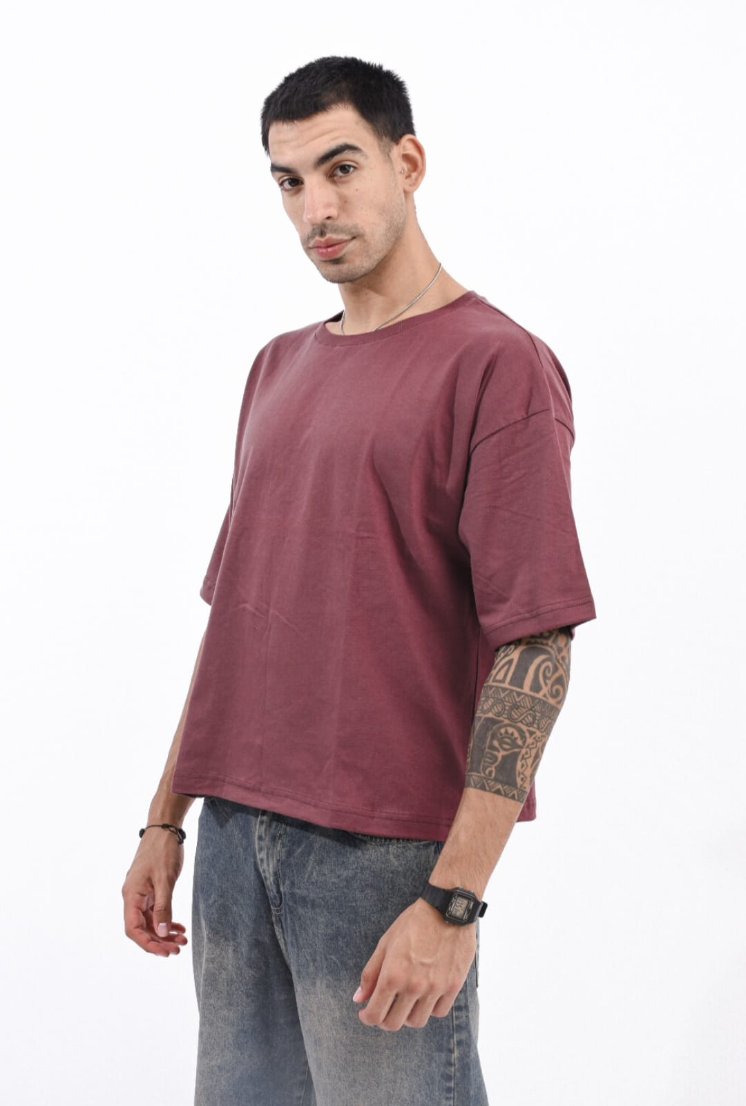 Hombre con camiseta extragrande color burdeos y vaqueros grises, de pie sobre un fondo blanco.