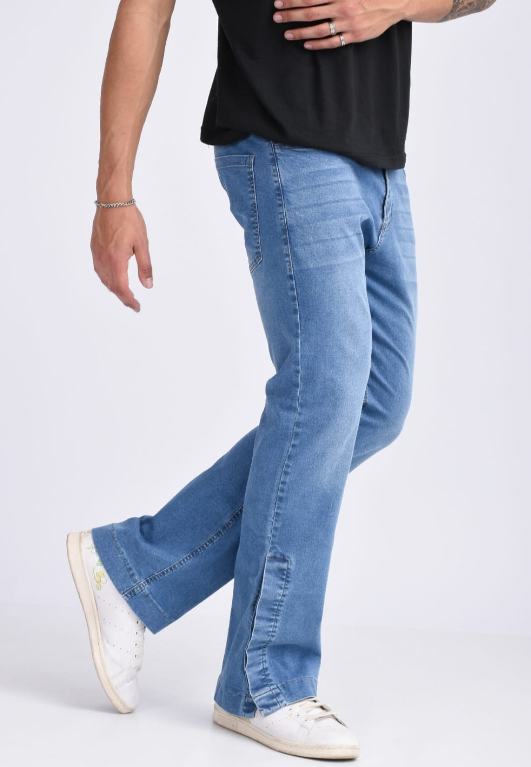Persona con camiseta negra, vaqueros azul claro y zapatillas blancas de pie sobre un fondo liso.