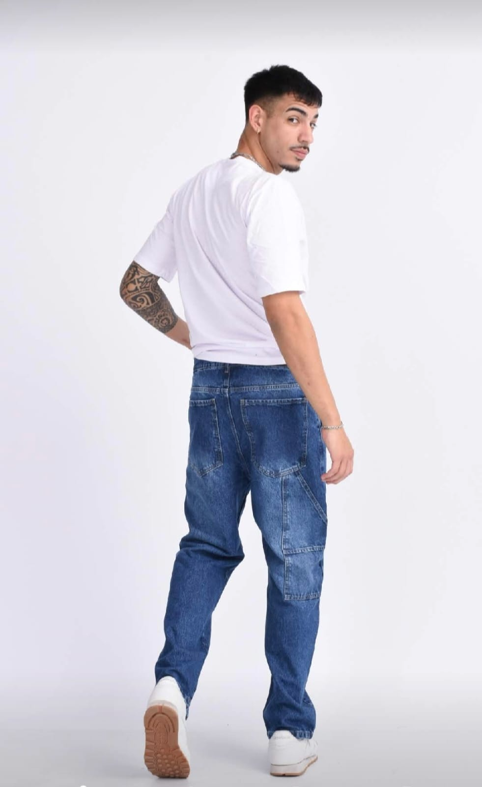 Hombre con camiseta blanca, pantalones negros, gorra y zapatillas blancas posando sobre un fondo gris claro.