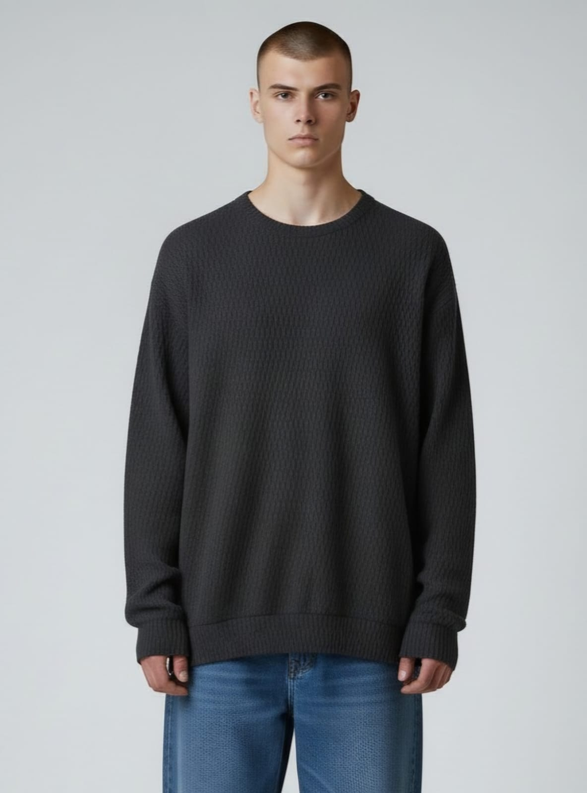 Persona de pie sobre un fondo gris, con un suéter oscuro extragrande y pantalones vaqueros azules.