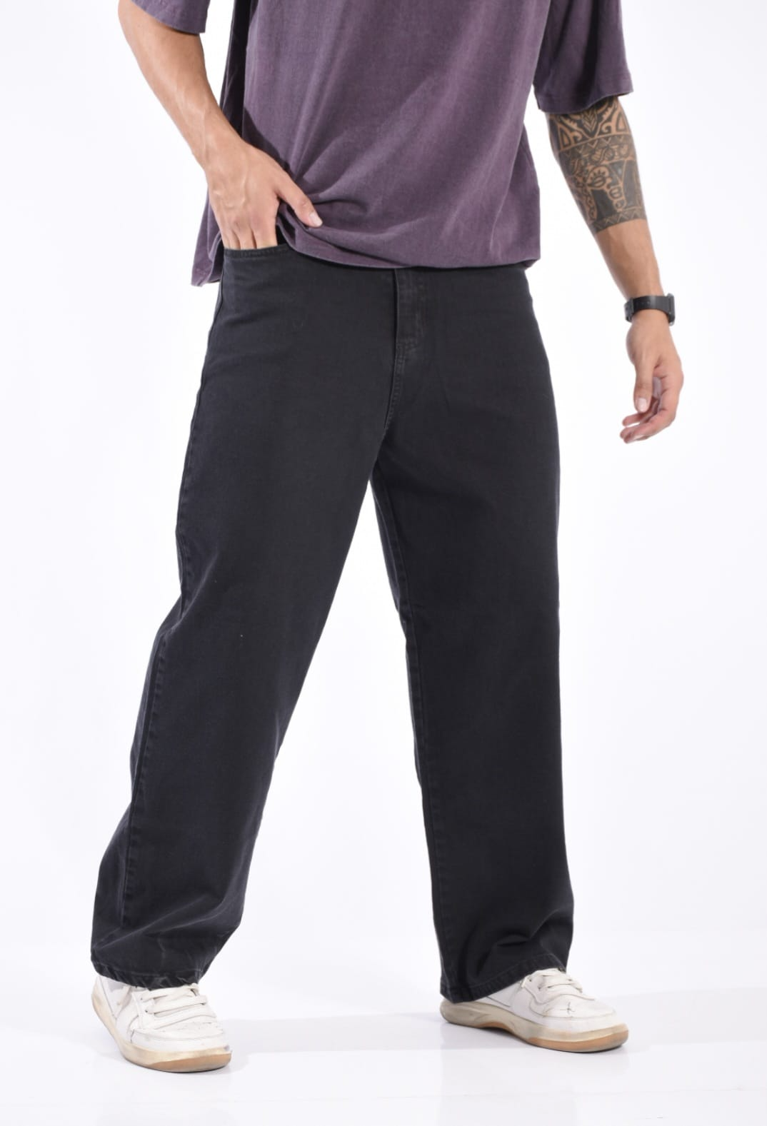 Persona con pantalones negros de pierna ancha, camisa morada y zapatillas blancas sobre un fondo blanco liso.