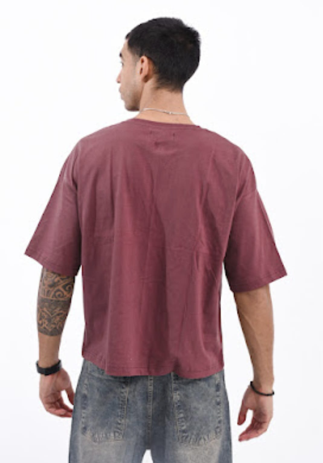 Hombre con camiseta extragrande color burdeos y vaqueros grises, de pie sobre un fondo blanco.