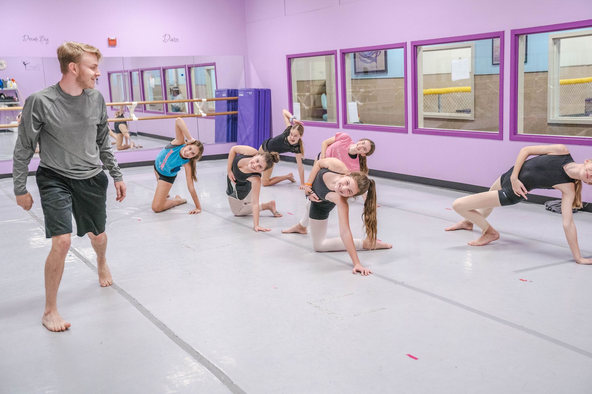 A man is standing next to a group of young girls doing exercises in a dance studio.