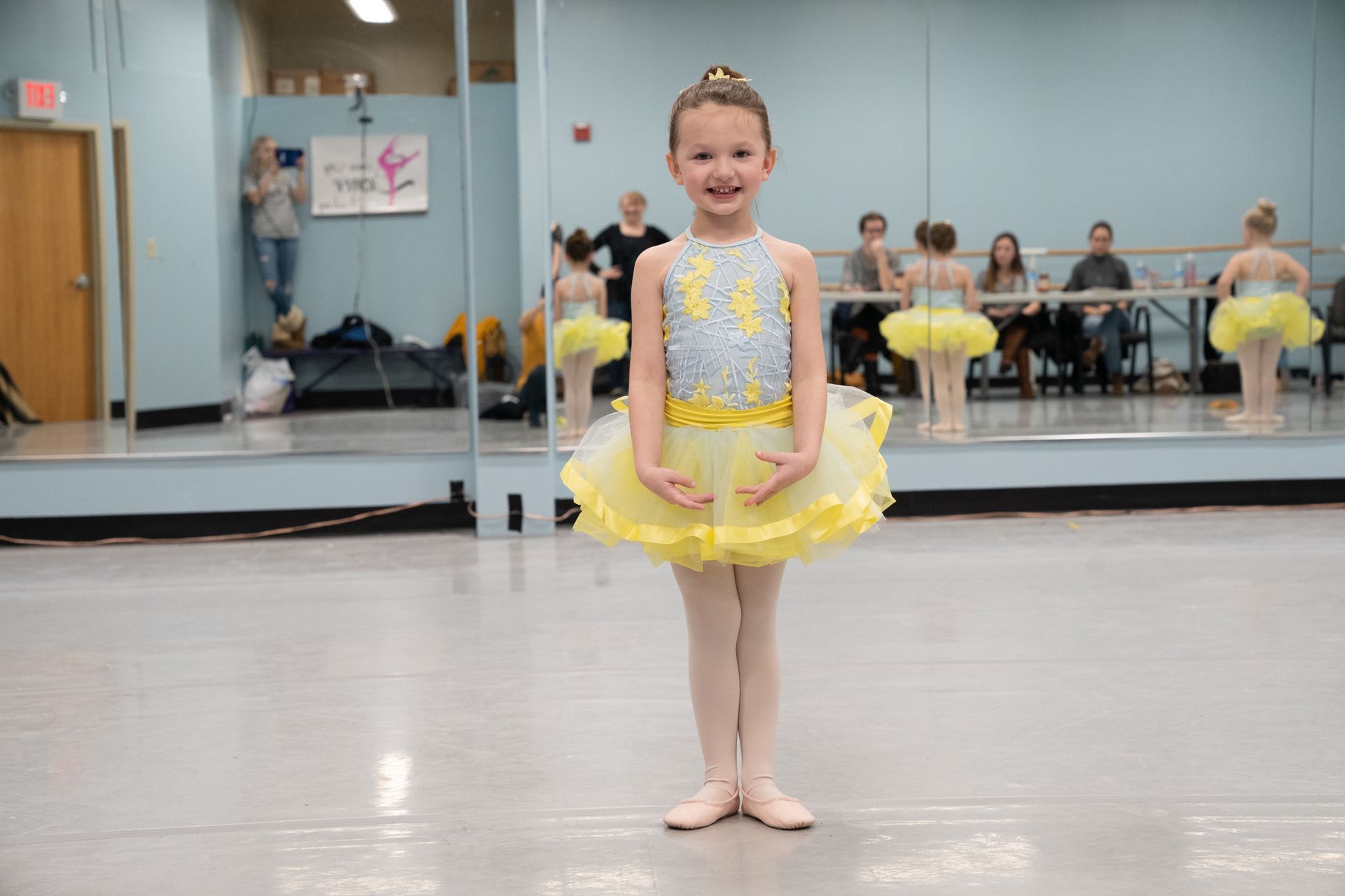 A little girl in a yellow tutu is standing in front of a mirror in a dance studio.