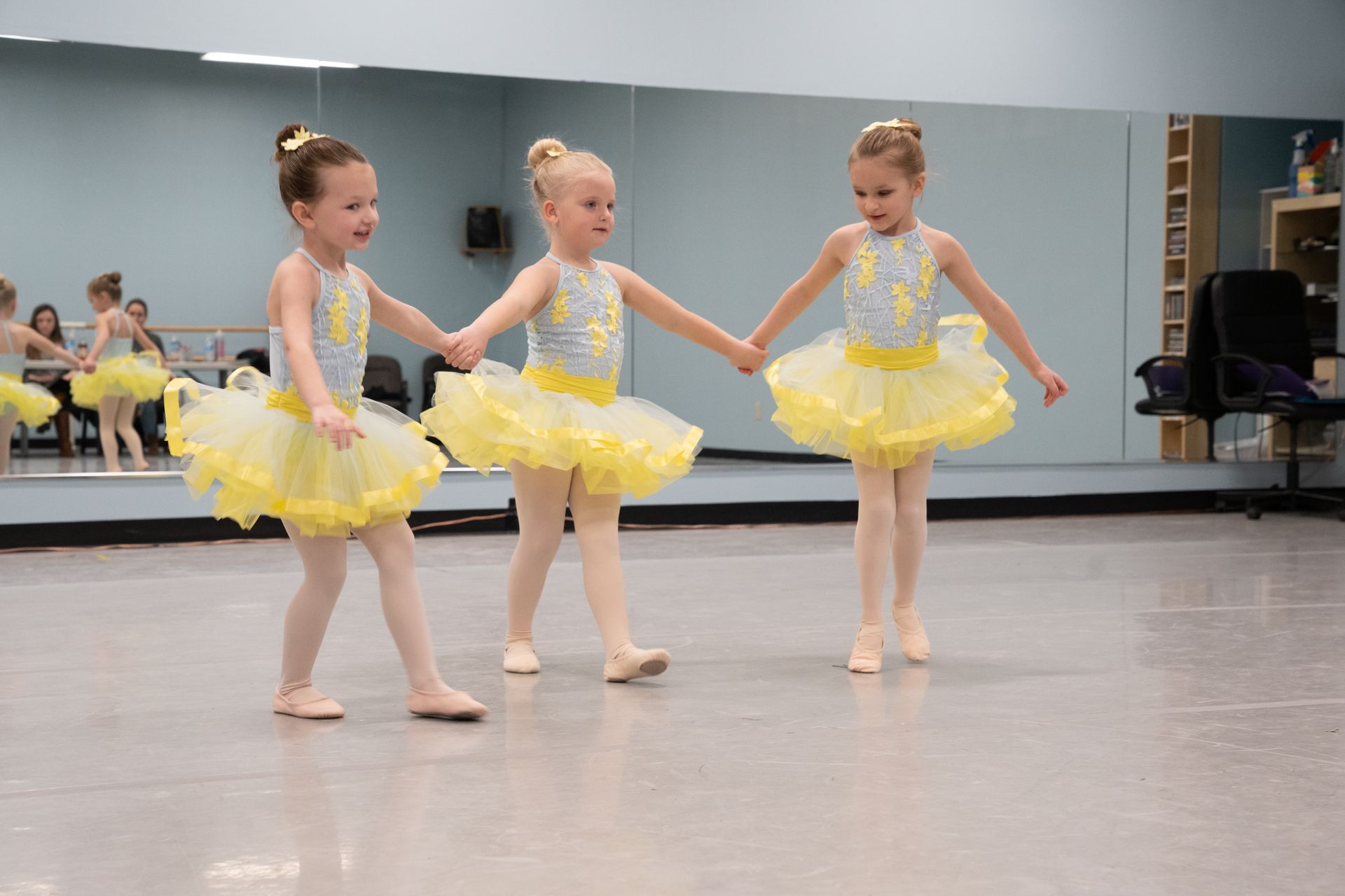 Three little girls in yellow tutus are holding hands in a dance studio.
