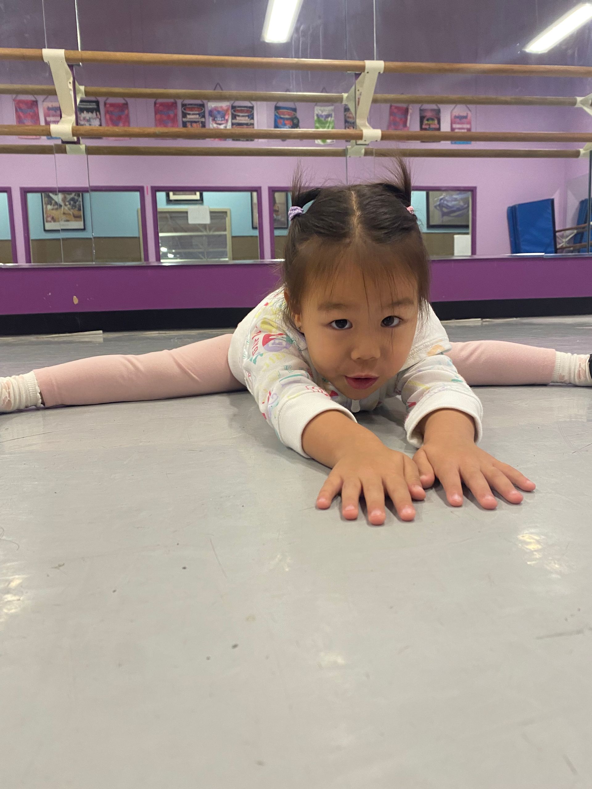 A little girl is doing a split on the floor in a dance studio.