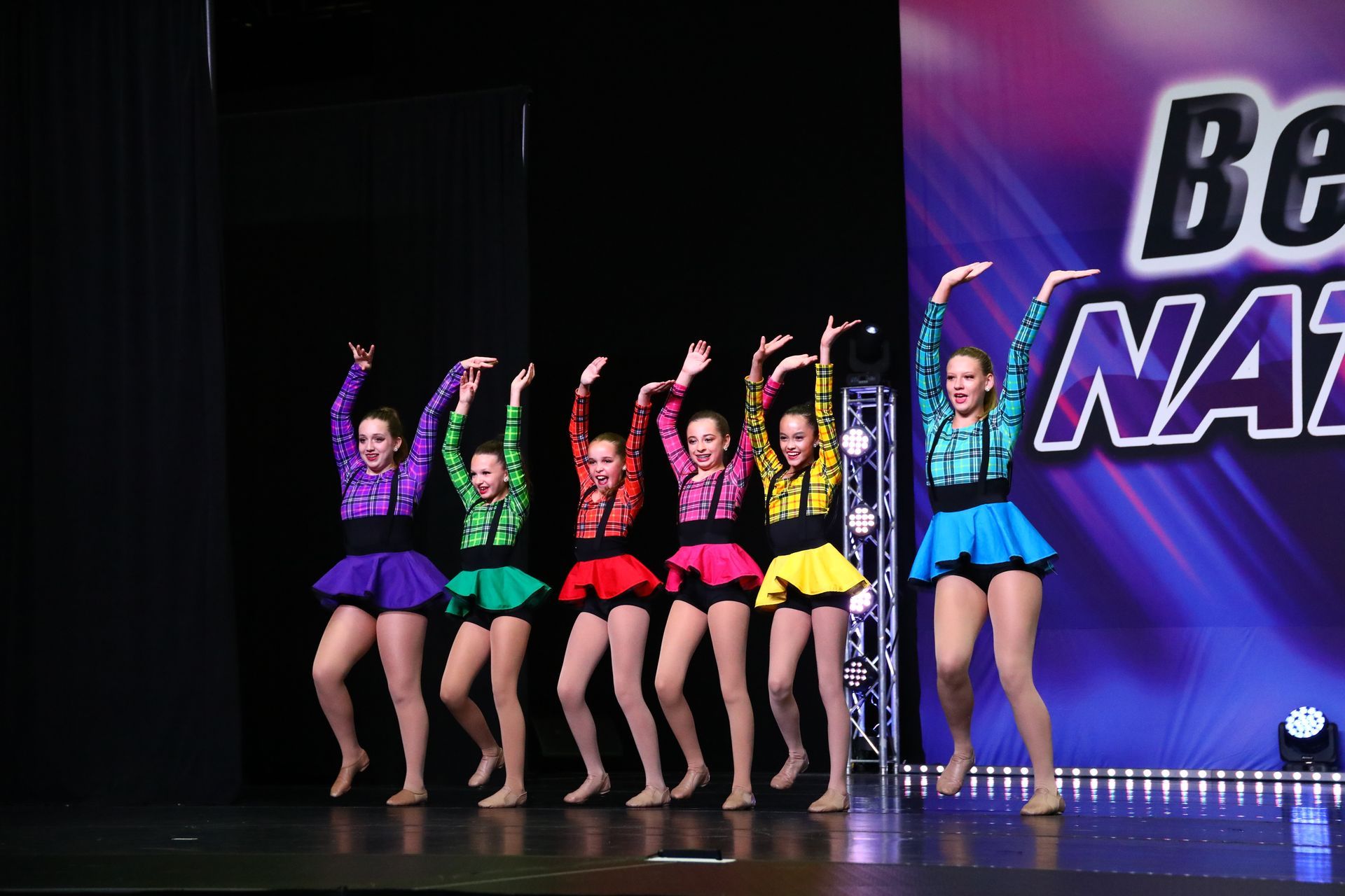 A group of young girls are dancing on a stage in front of a sign that says be national