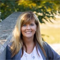 Woman with blonde hair, smiling, wearing a blue jacket and white shirt outdoors.