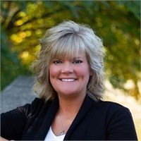 Blonde woman smiling, wearing a black blazer. Outdoor setting with blurred background.
