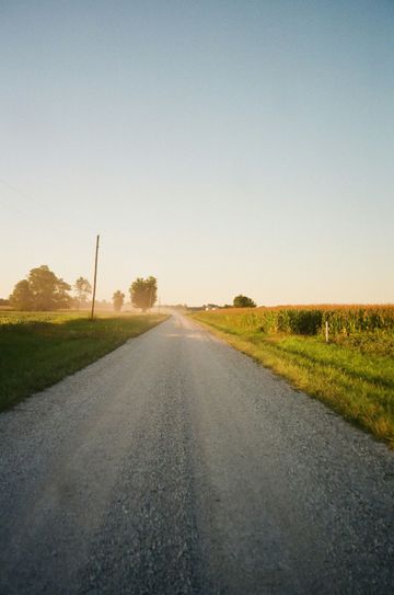 Gravel road leading into the distance, flanked by green fields and trees, under a pale blue sky.