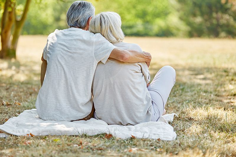 Two people sit on a blanket in a sunny park, embracing and looking out over the grass.