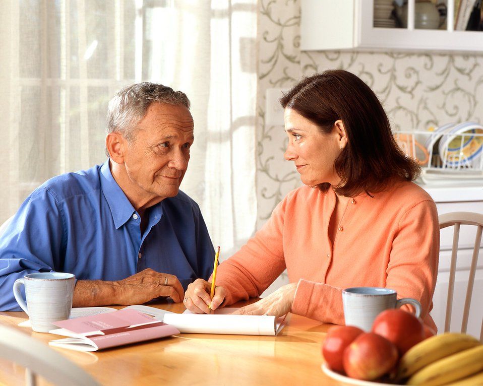 Two people talking at a kitchen table with mugs, fruit, and papers in a bright home kitchen.