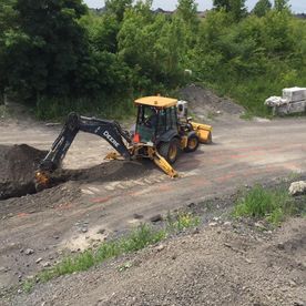 Une pelle mécanique creuse une tranchée dans un terrain gravillonné, avec des arbres en arrière-plan. Matériel John Deere.