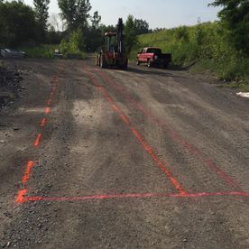 Des bandes de peinture orange en aérosol délimitent une zone triangulaire sur un chemin de gravier ; des engins de chantier sont visibles en arrière-plan.