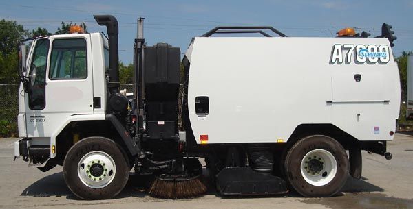 Camion balayeuse de rue blanche avec brosses latérales et un grand tuyau d'échappement, stationné à l'extérieur.
