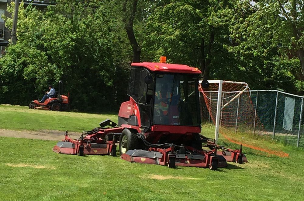 Tondeuse commerciale rouge sur un champ vert ; une autre tondeuse à l’arrière-plan. Un but de football est visible.