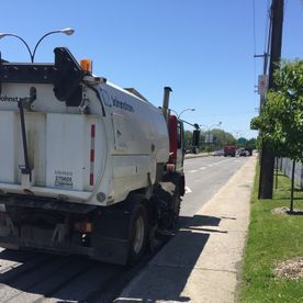 Balayeuse de rue nettoyant la chaussée ; journée ensoleillée, vue depuis le trottoir.