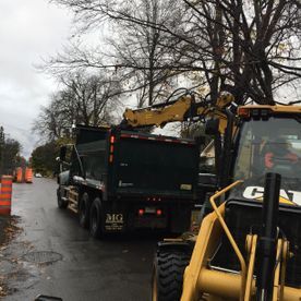 Une pelleteuse jaune charge un camion-benne noir sur une route mouillée. Des cônes de signalisation orange bordent la rue.