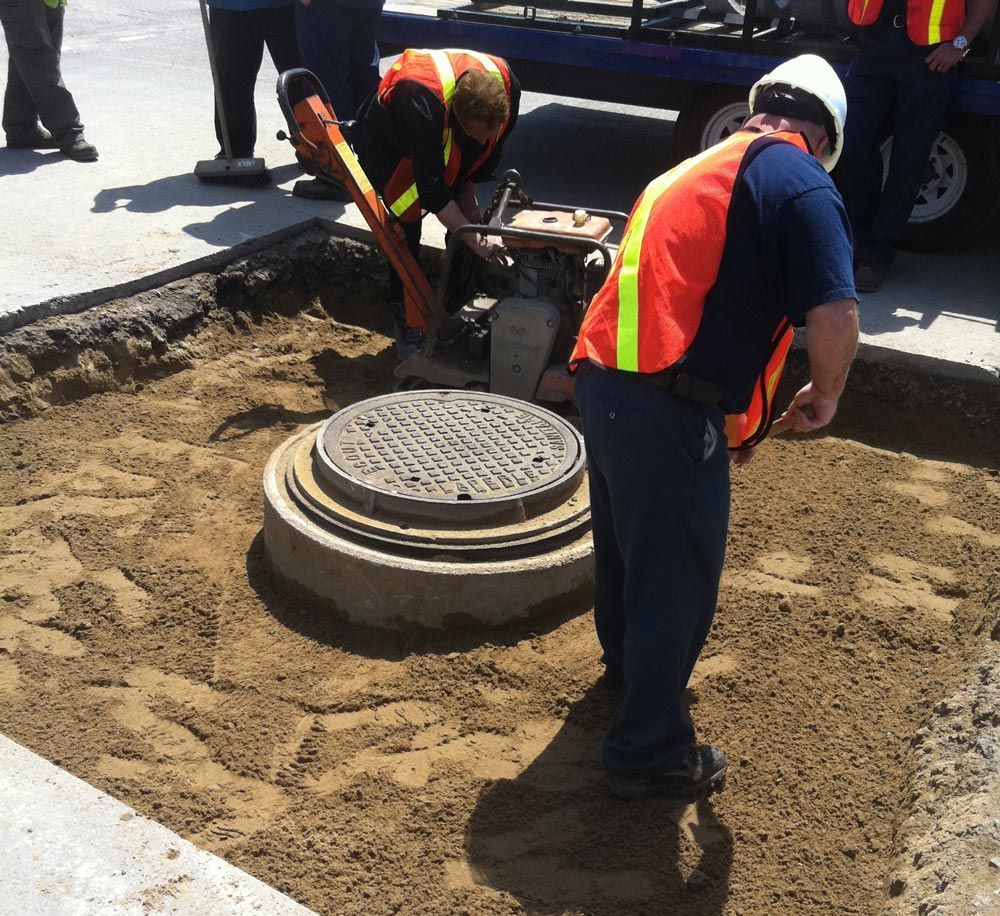 Des ouvriers portant des gilets de sécurité réparent une bouche d'égout au niveau de la rue, à l'aide d'outils et de machines.