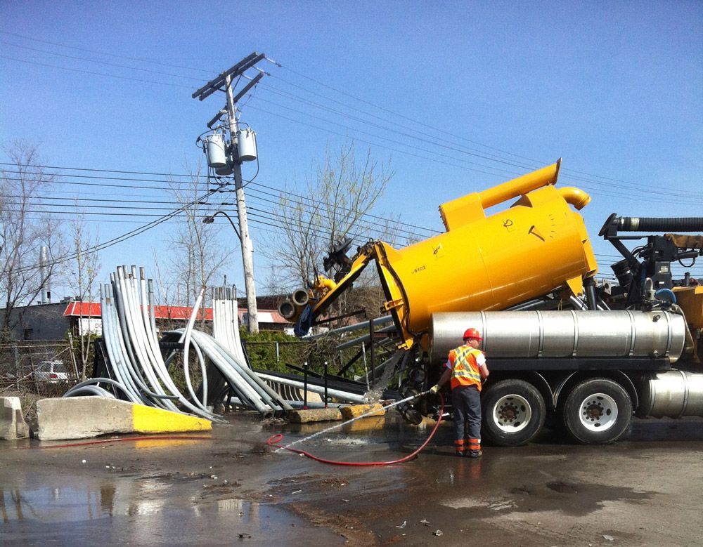 Un camion-citerne jaune déverse des débris. Un ouvrier en gilet orange arrose les structures métalliques endommagées sous un poteau électrique.