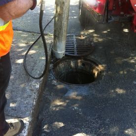 Une personne portant un gilet de sécurité nettoie une canalisation avec un tuyau d'arrosage sur une surface en béton.