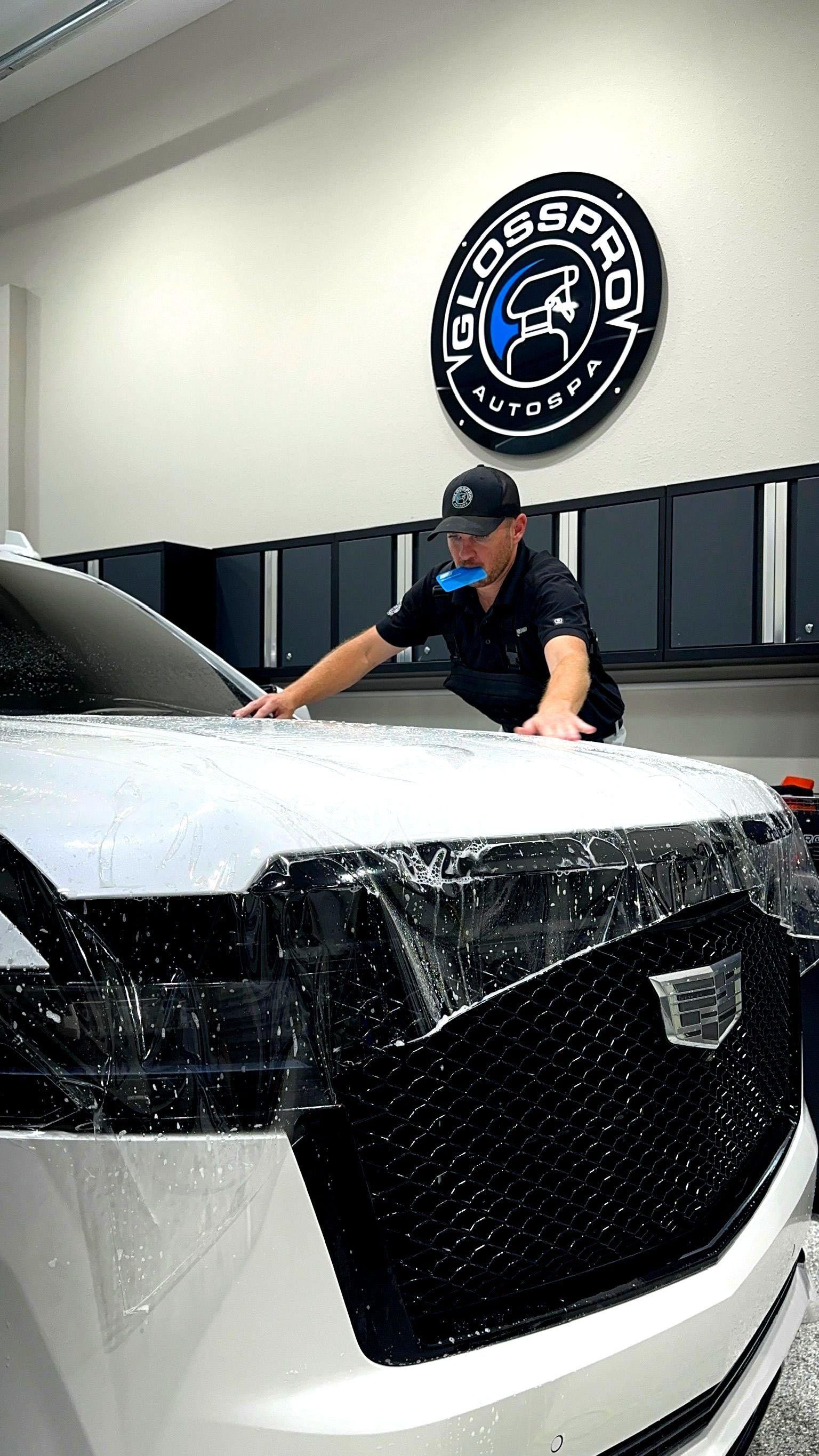 A person applies protective film to the front of a white Cadillac in a shop with the 