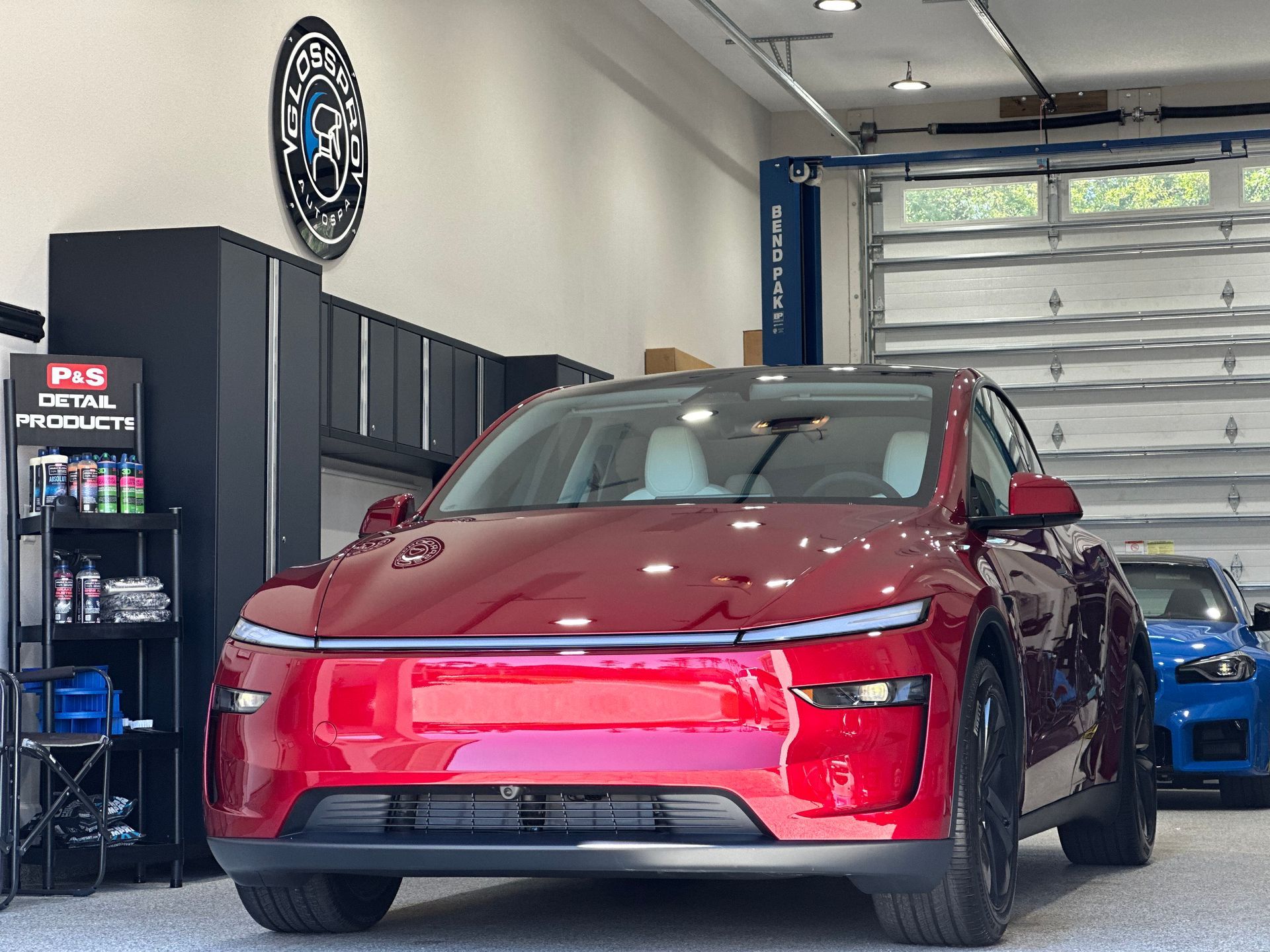A red electric car parked in a garage. The car is in front of open garage doors, and a blue car is visible to its right.